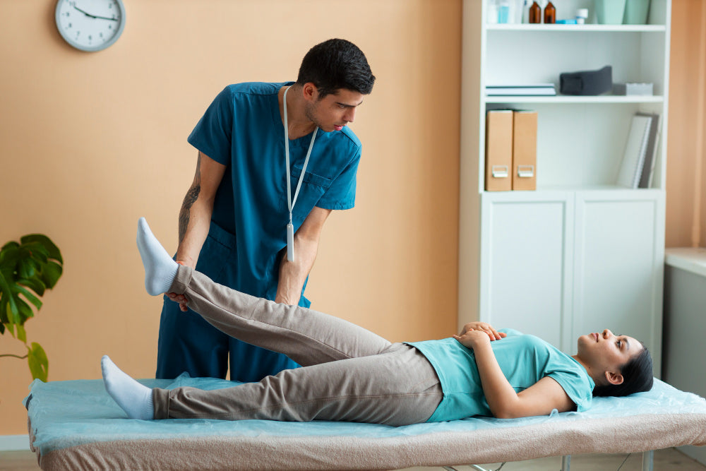 Healthcare professional in teal scrubs assisting a patient with a leg rehabilitation exercise on a treatment table in a physio center in Dubai.