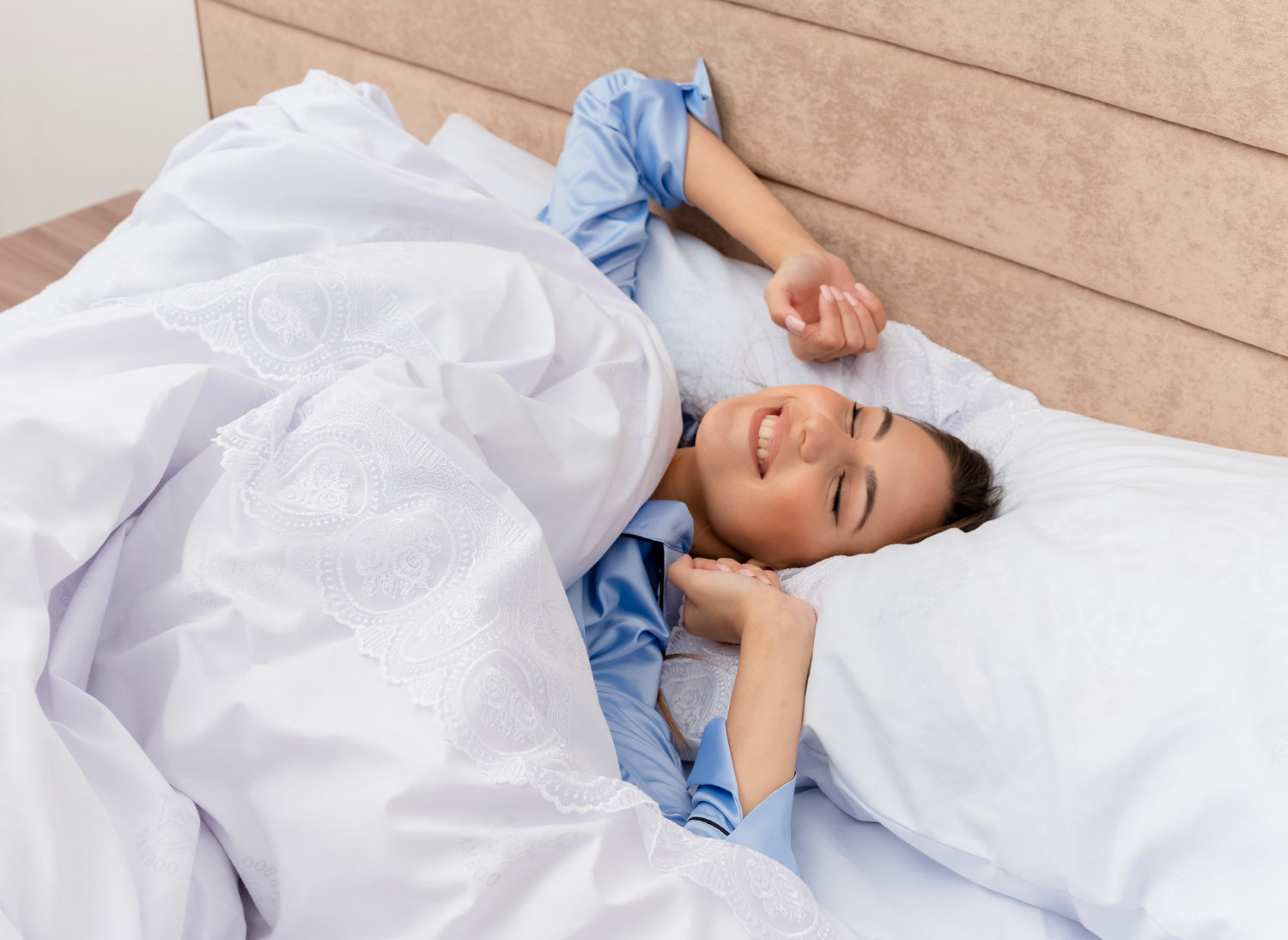 A woman smiling and stretching while lying on her white bed, demonstrating a comfortable sleeping position for back pain relief